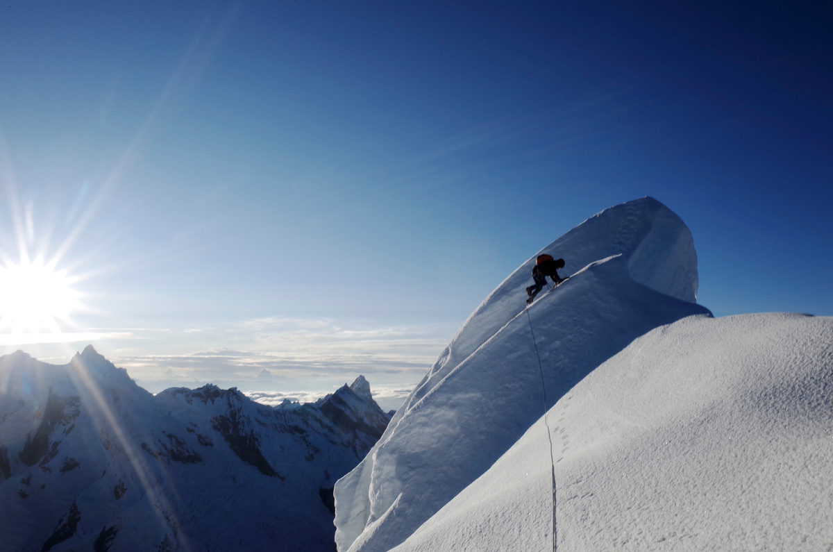 A climber on a corniced snow ridge with the sun bursting over the peaks behind. Alpamayo summit in the Cordillera Blanca