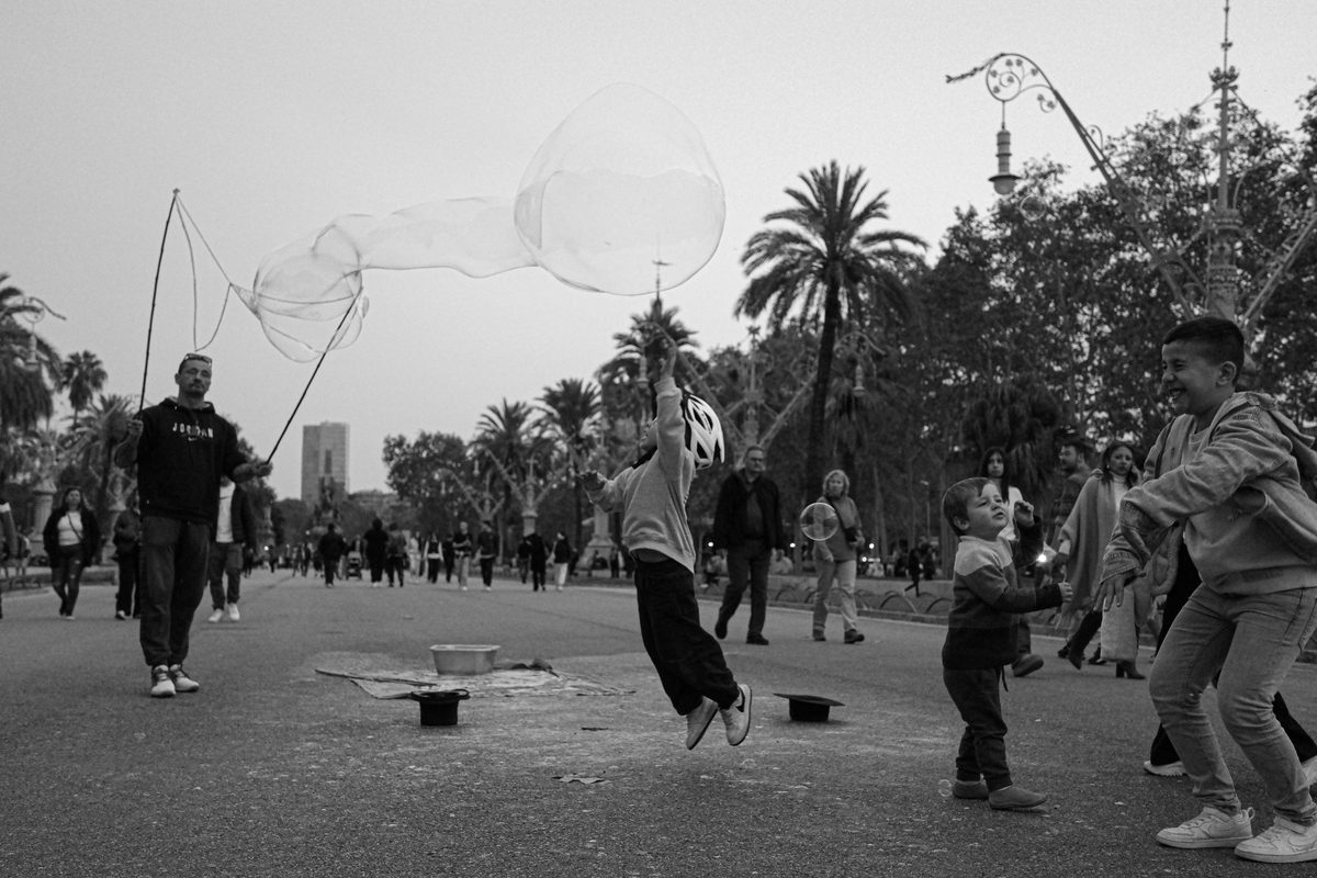 Black and white street scene of children chasing giant soap bubbles along a palm-lined plaza