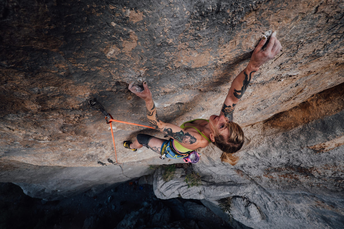 A climber in a yellow top reaching high on an overhanging wall, the rope running down to a quickdraw