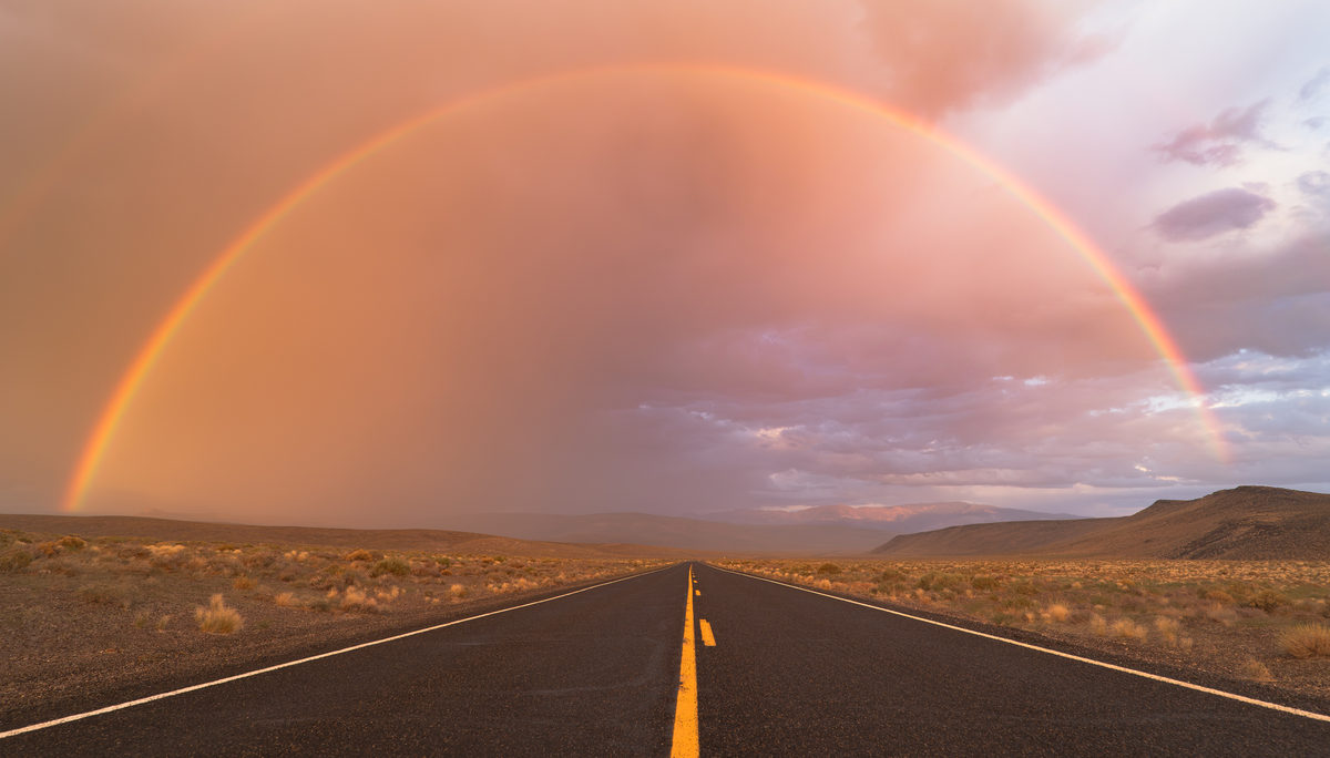 A full rainbow arcs over an empty desert highway at sunset